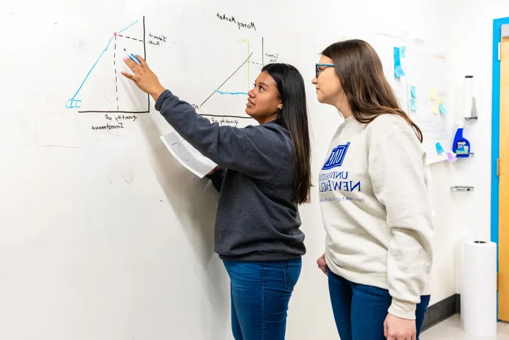 A U N E student explains a graph drawn on a whiteboard, titled "Money Market," to a fellow student